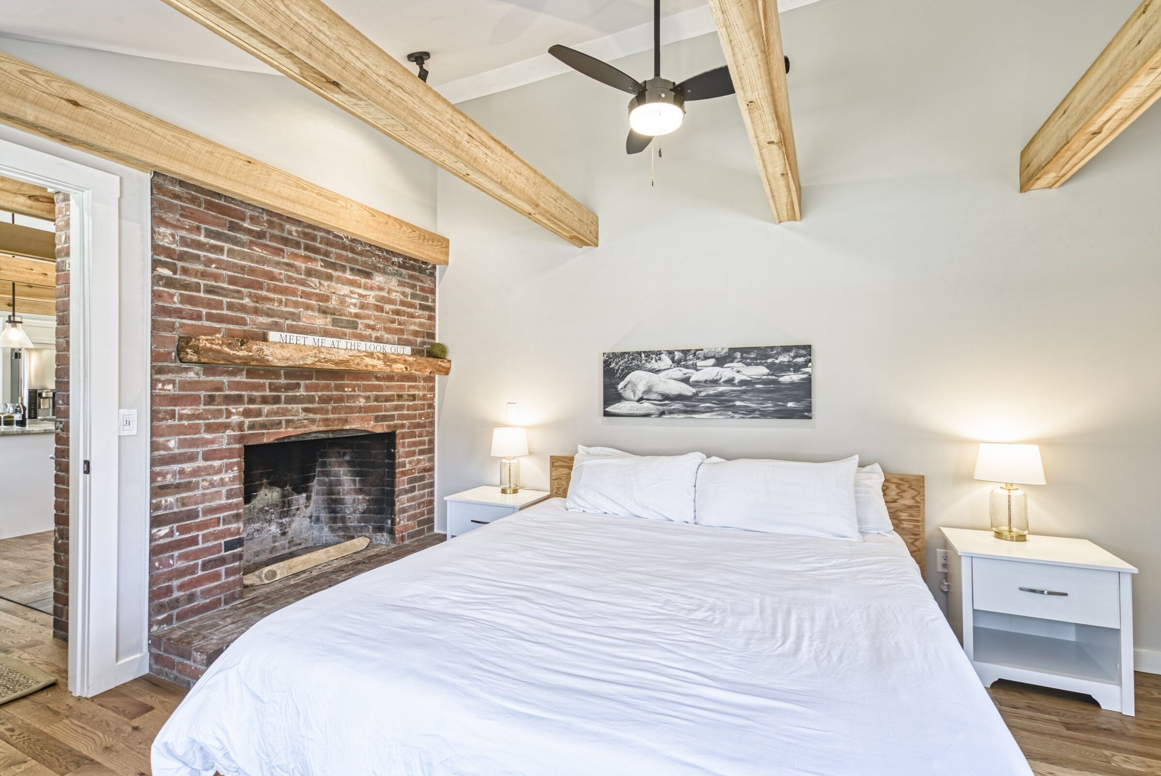 A bedroom with a brick fireplace, wooden beams, a ceiling fan, white bedding, and bedside lamps.