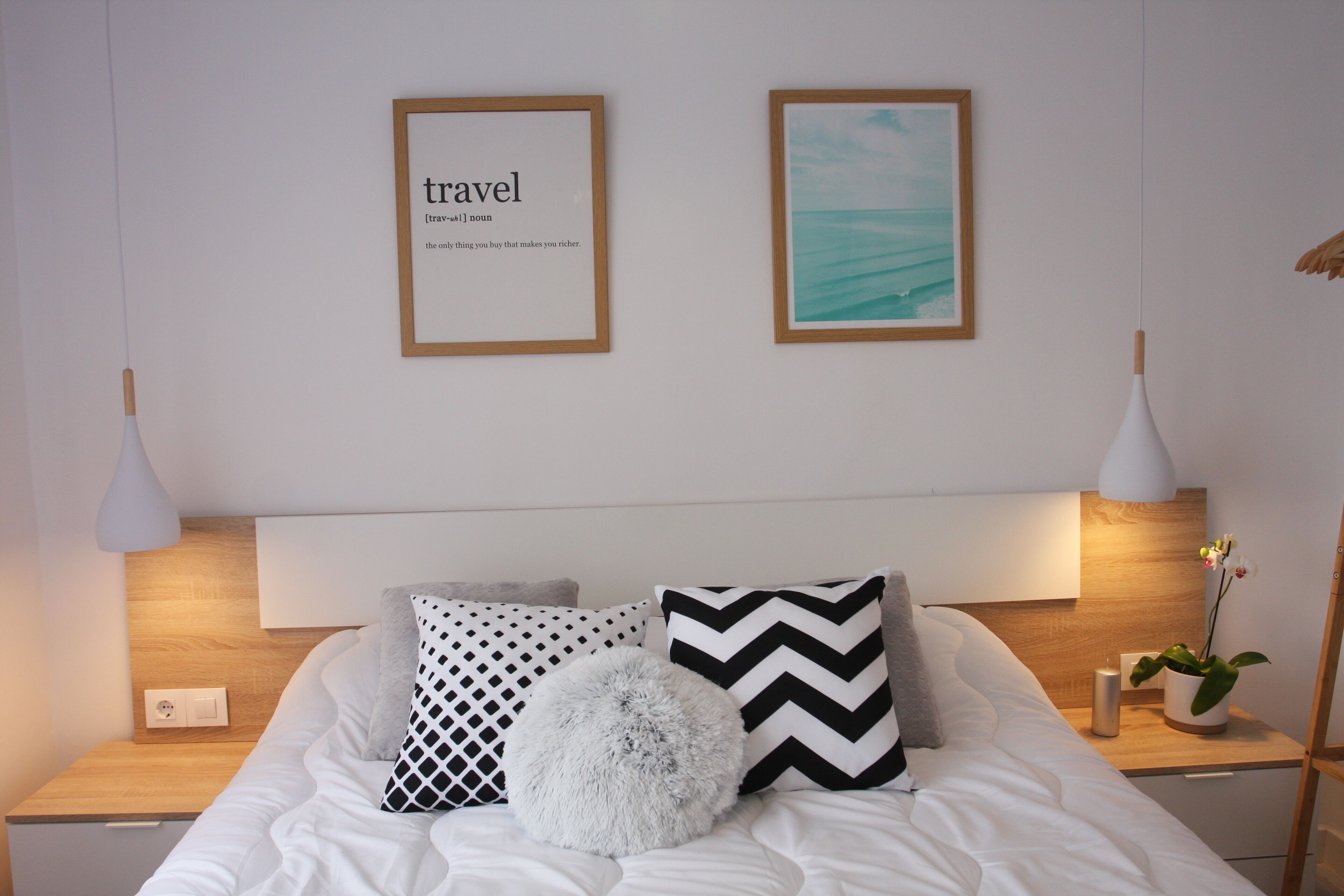 Un lit bien fait, recouvert d’une couette blanche et agrémenté de plusieurs oreillers, dont un moelleux et deux ornés de motifs géométriques. On y trouve deux tables de chevet avec des lampes, et un tableau encadré est accroché au mur au-dessus du lit.