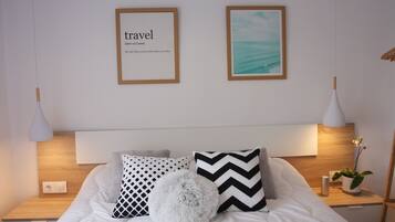 Un lit bien fait, recouvert d’une couette blanche et agrémenté de plusieurs oreillers, dont un moelleux et deux ornés de motifs géométriques. On y trouve deux tables de chevet avec des lampes, et un tableau encadré est accroché au mur au-dessus du lit.