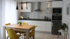 A modern kitchen with white cabinets, a stainless steel refrigerator, and a microwave. There is a dining table with yellow chairs and a potted plant on the side. - Edificio Privado La Herradura (Haro)