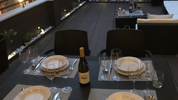 An outdoor dining area with tables set for two, each with plates, cutlery, and wine glasses. There are potted plants along the railing and a view of buildings in the background. - Edificio Privado La Herradura (Haro)