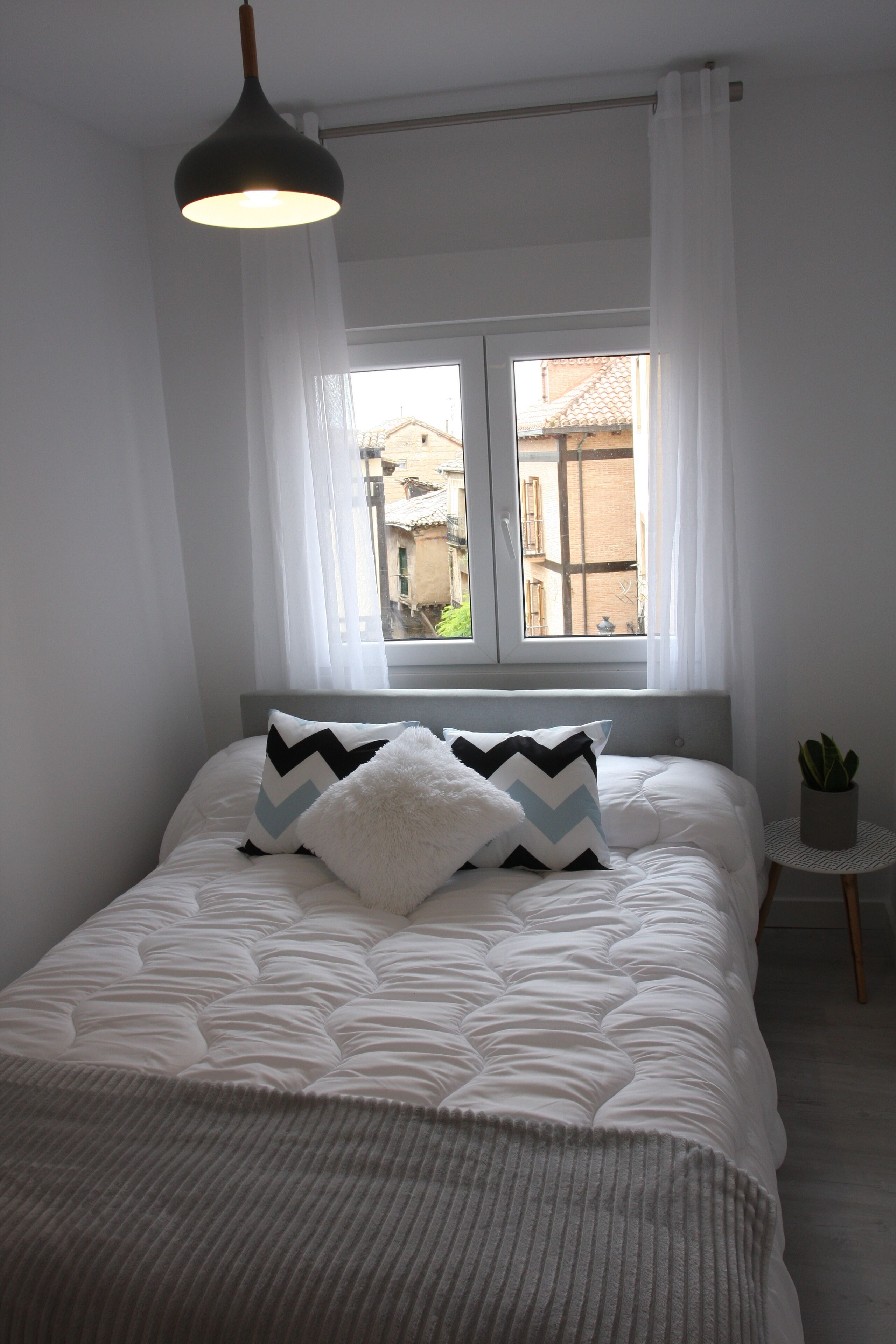 A neatly made bed with a white duvet and two pillows, a small potted plant, and a window with white curtains.
