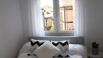A neatly made bed with a white duvet and two pillows, a small potted plant, and a window with white curtains.
