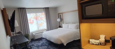 Une chambre d’hôtel comprenant un lit, un bureau, une chaise, une lampe de bureau, une fenêtre avec des rideaux, un four à micro-ondes et une machine à café.