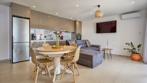Een moderne keuken met een eethoek, een grijze bank en een aan de muur bevestigde televisie.