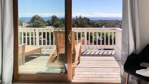 Un balcon doté d’une terrasse en bois, d’une balustrade blanche et offrant une vue sur l’océan et les arbres.