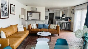 Un salon avec un canapé marron, une table basse blanche et un fauteuil bleu. Au fond, on aperçoit une cuisine avec un réfrigérateur et un évier.