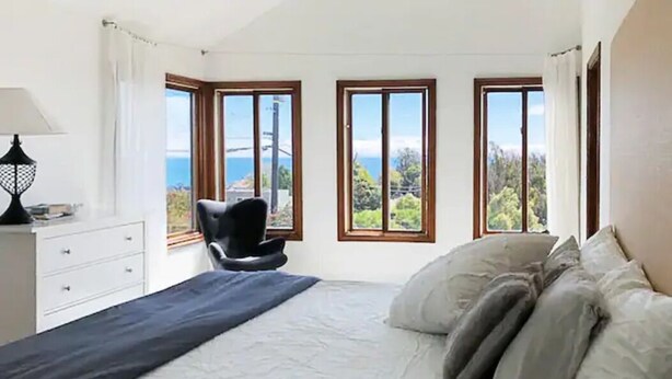 Une chambre à coucher comprenant un lit, une chaise et trois fenêtres donnant sur la mer et la verdure.