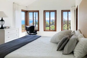 Une chambre à coucher comprenant un lit, une chaise et trois fenêtres donnant sur la mer et la verdure.