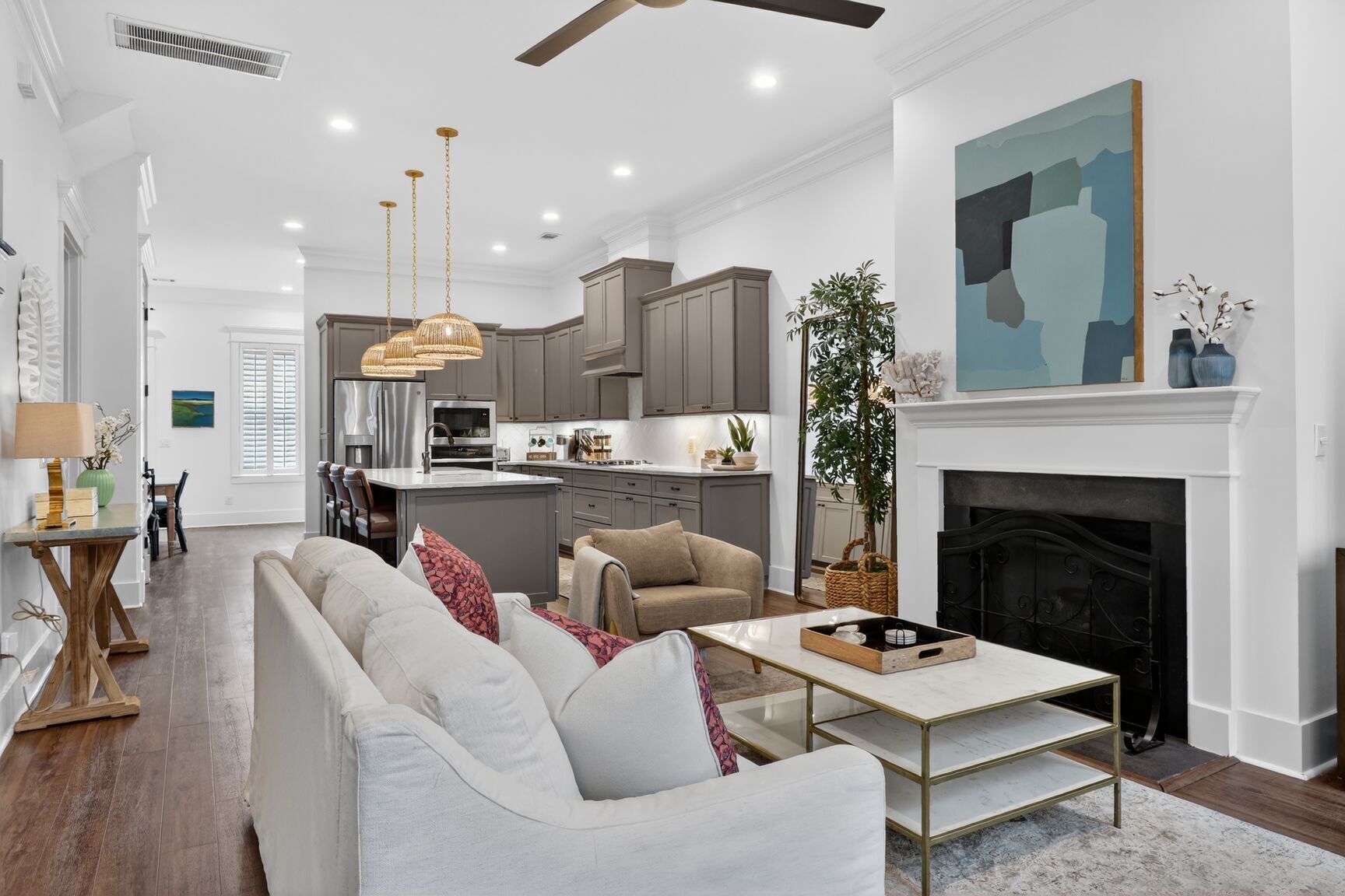 A spacious living room with a sofa, coffee table, and fireplace. A kitchen with grey cabinets and a dining area with a table and chairs are visible in the background.