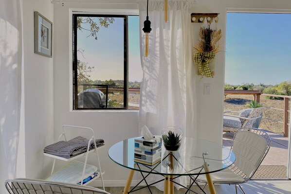 Ein Zimmer mit einem Glastisch, zwei Stühlen, einer kleinen Topfpflanze und einem Fenster mit Blick ins Freie.