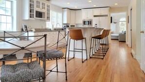 Una cocina amplia con armarios blancos, una isla central con taburetes de bar, una lámpara colgante y suelo de madera.