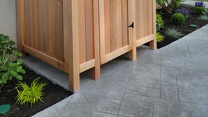 Un cobertizo exterior de madera con dos puertas y un cierre, ubicado en un patio empedrado con un jardín al fondo.