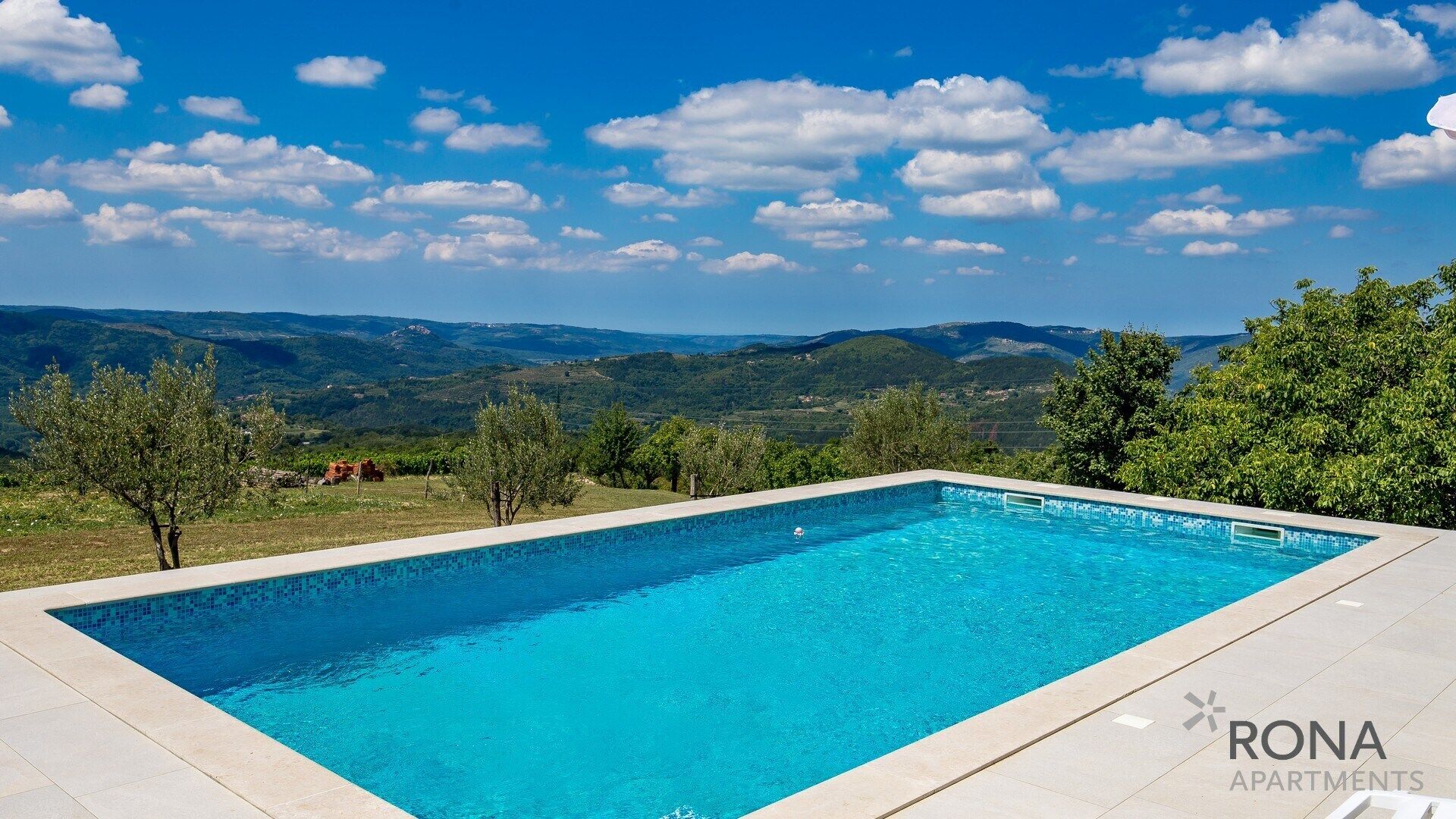 A rectangular swimming pool with clear blue water, surrounded by a tiled deck, set against a backdrop of green hills and a clear blue sky with scattered clouds.