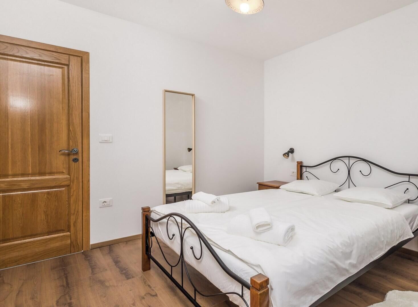 A bedroom with a wooden door, a bed with a metal frame, two bedside tables, a mirror, and a small desk with a chair.