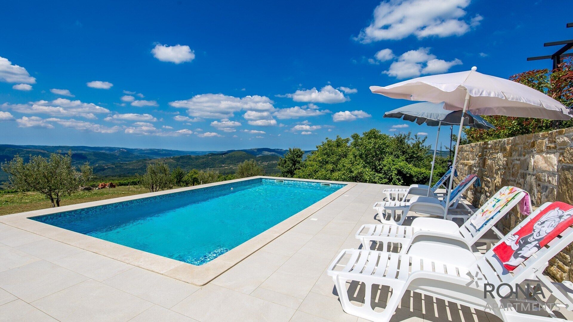 Une piscine rectangulaire à l’eau d’un bleu cristallin, entourée de chaises longues blanches et d’un mur en pierre.