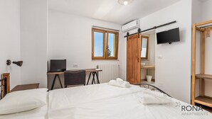 A bedroom with a bed, a desk with a computer, a window, a mirror, and a TV mounted on the wall.