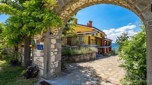 A stone archway leading to a yellow house with a stone wall and greenery on the left.