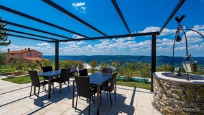 Eine Terrassenanlage im Freien mit einer Pergola, einem Tisch mit Stühlen, einer Steinmauer und Blick auf ein Haus und Berge.