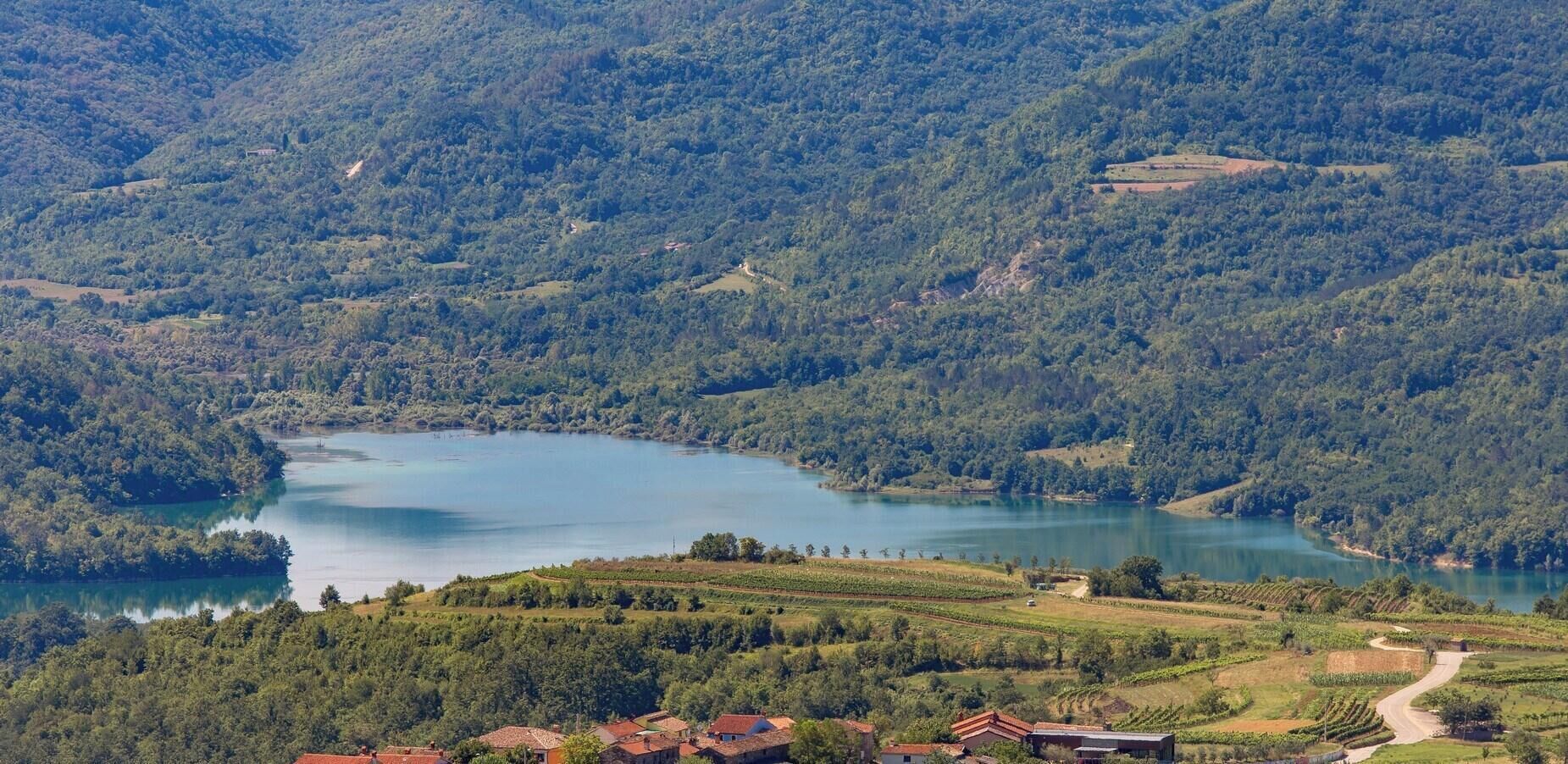 A scenic view of a lake surrounded by lush greenery and terraced vineyards, with traditional buildings in the foreground.