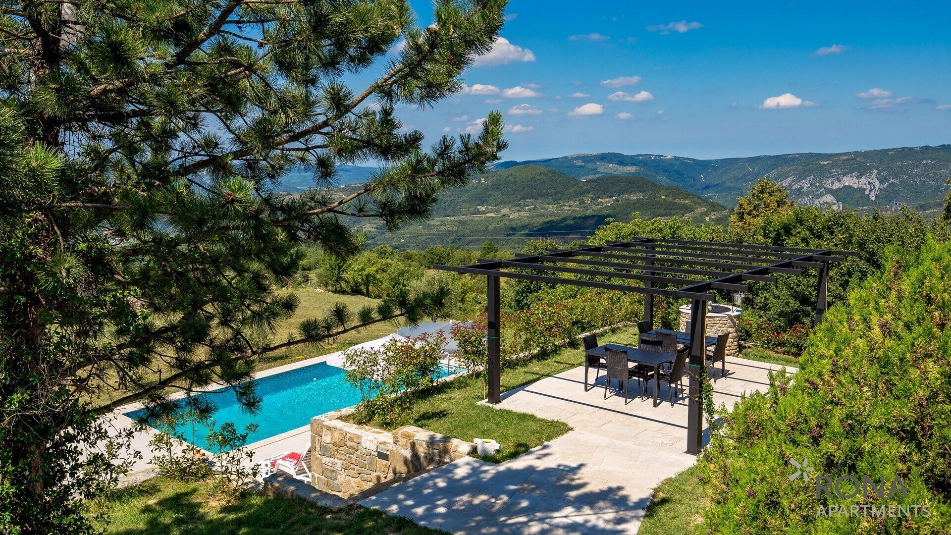 Una piscina con un pergolato e posti a sedere esterni, circondata da una vegetazione lussureggiante e uno sfondo montuoso.