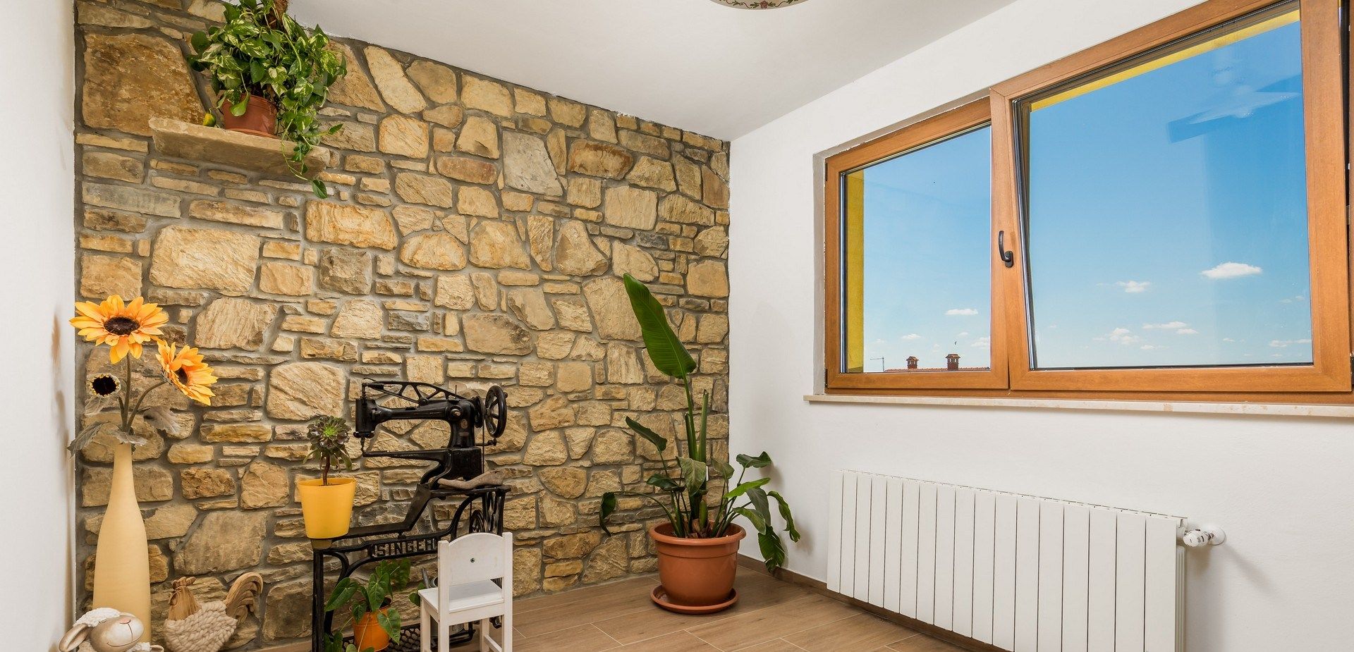 A room with a stone wall, a sewing machine, a chair, a radiator, and a window with a view of the sky.
