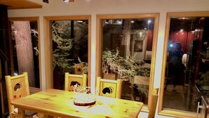 Zona de menjador amb taula i caires de fusta, grans finestres que donen vistes a arbres i un edifici.