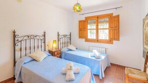 Ein Schlafzimmer mit zwei Betten, einem Nachttisch, einer Lampe, einem Fenster mit Holzjalousien und einem Sessel.