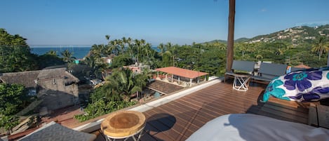 A wooden deck with a thatched roof, a bed with a floral pillow, and a view of the ocean and hills.