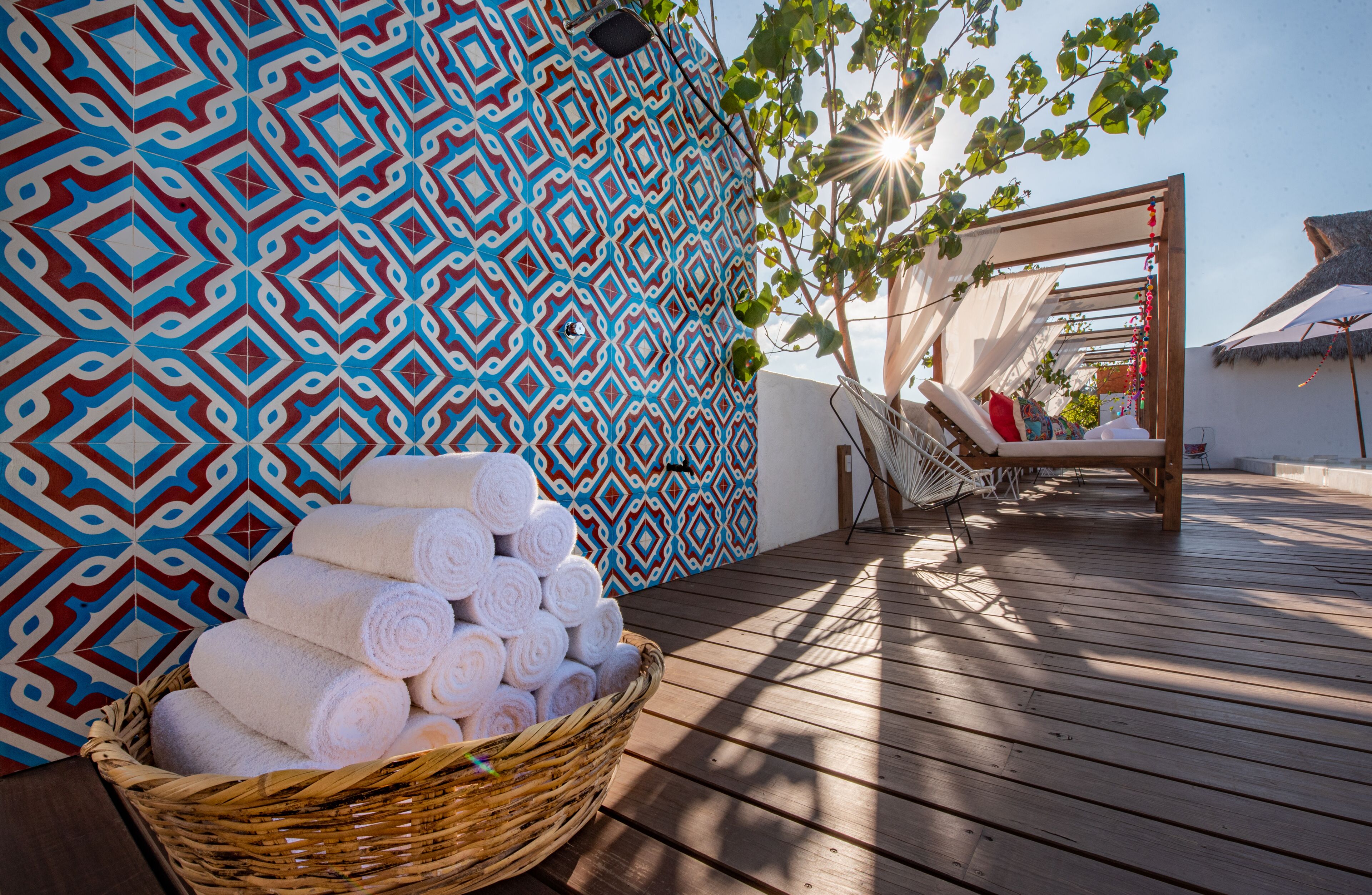 A wooden deck with a patterned wall, a wicker basket of towels, and a sunlit relaxation area with a hammock and lounge chairs.