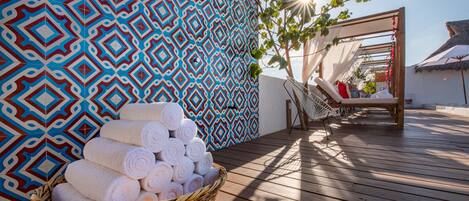 A wooden deck with a patterned wall, a wicker basket of towels, and a sunlit relaxation area with a hammock and lounge chairs.