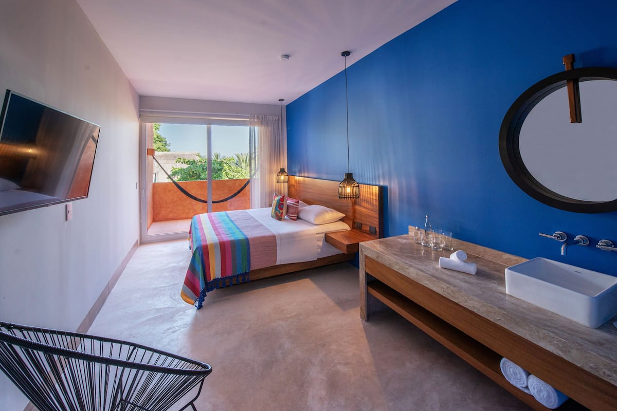 A modern hotel room with a bed, a wooden vanity with a sink, a large mirror, and a television mounted on the wall.