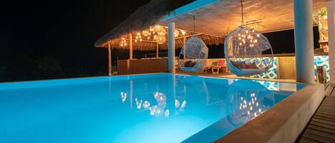 A swimming pool with hanging chairs and a thatched-roof structure.