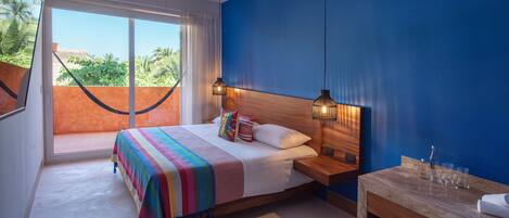 A bedroom with a large bed, a wooden nightstand, a small table with glasses, and a view of greenery through a window.