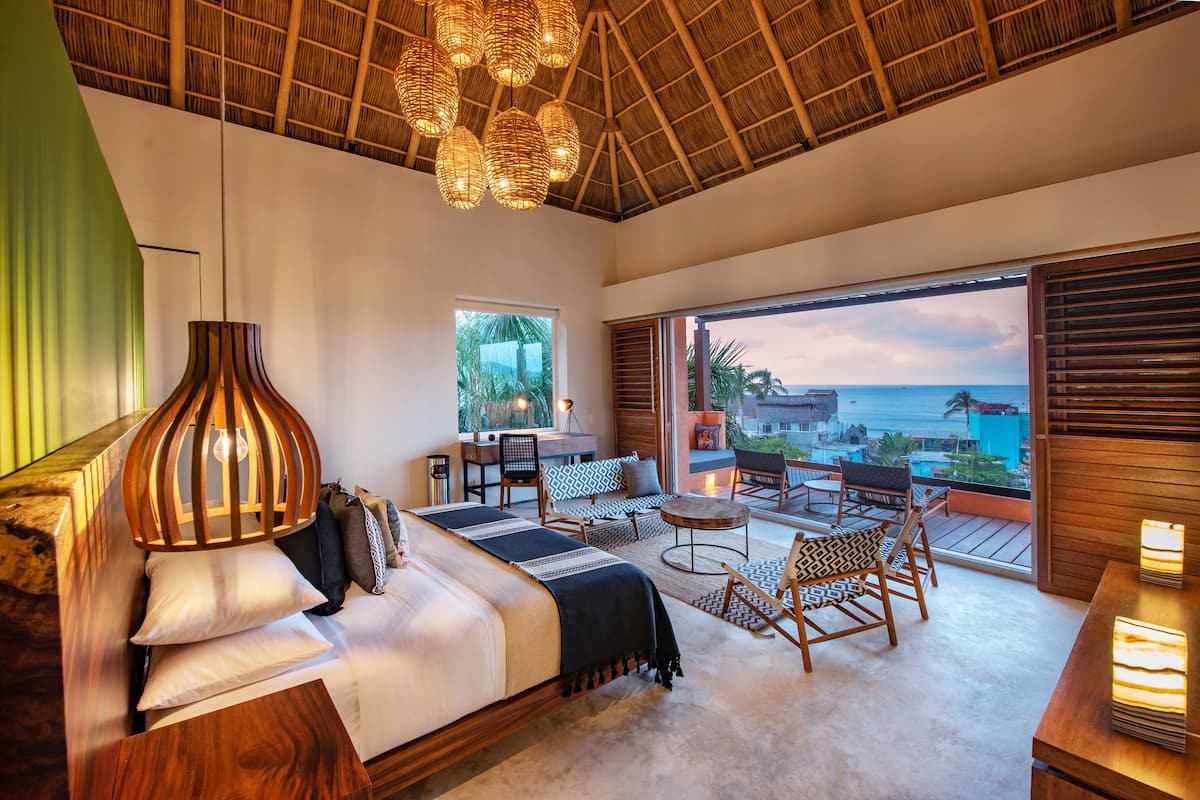 A bedroom with a bed, a seating area with chairs and a table, and a view of the ocean.