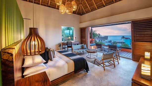 A bedroom with a bed, a seating area with chairs and a table, and a view of the ocean.