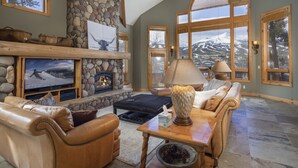 A spacious living room with a stone fireplace, leather sofas, and large windows offering mountain views. - Walking distance to downtown Breck, Stunning ski resort panorama, hot tub, foosball, arcade machine! (Breckenridge)