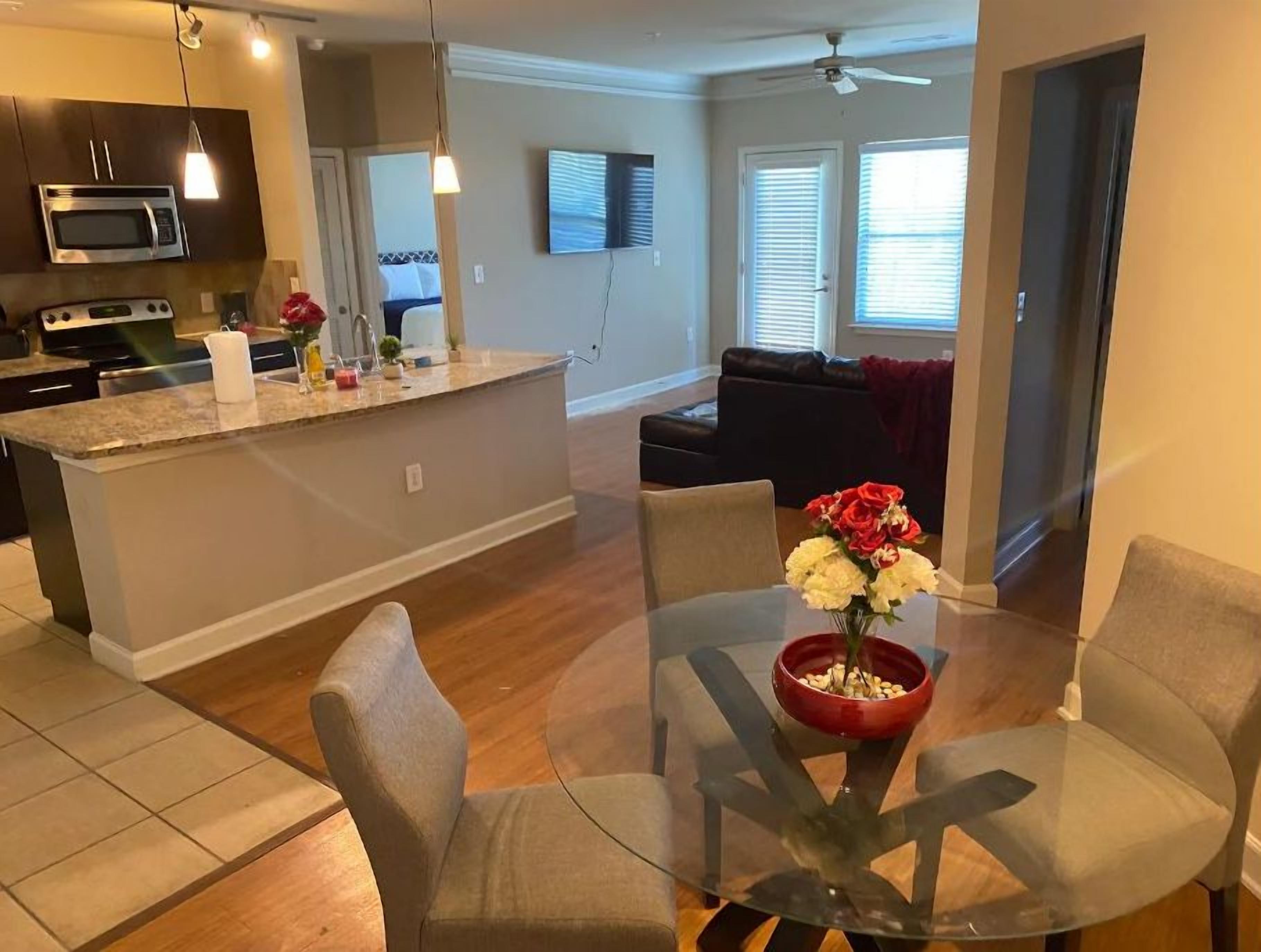A modern kitchen with a central island, a dining area with a glass table and chairs, and a living room with a sofa and a TV.