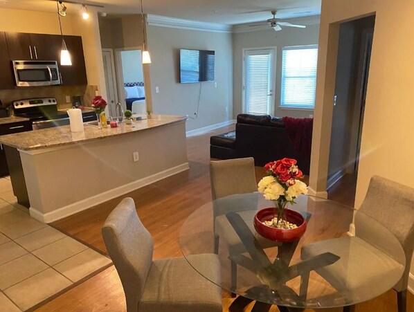 A modern kitchen with a central island, a dining area with a glass table and chairs, and a living room with a sofa and a TV.