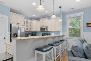 A kitchen with white cabinets, a stainless steel refrigerator, a microwave, and a stove. There are three bar stools and a window with blinds.