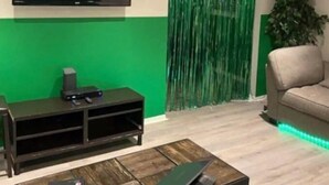 A living room with a flat-screen TV mounted on the wall, a dark wooden coffee table, a beige sofa, and a green curtain with vertical lines. - Sale!!! Emerald Grande family home (Atlanta)