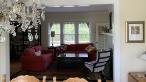 Ein Wohnzimmer mit einem roten Sofa, einem schwarzen Couchtisch und einer Kronleuchter. Im Hintergrund steht ein Bücherregal und es gibt ein Fenster mit Jalousien.