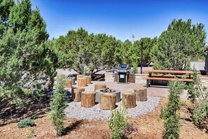 An outdoor patio area with a grill, wooden stools, and a picnic table surrounded by trees.
