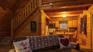 Interior acollidor d'una cabana de fusta amb una escala de fusta, un sofà amb coixins estampats, una cuina amb armaris de fusta i una taula de dinar.