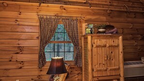 Interior acollidor d'una cabana amb parets de fusta, un espai de dormir a la llera alta, una llar de foc i una finestra amb cortines.