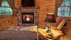 Interior acollidor d'una cabana amb una llar de pedra, un televisor penjat a sobre i una zona de seients confortable amb una taula i una làmpada.