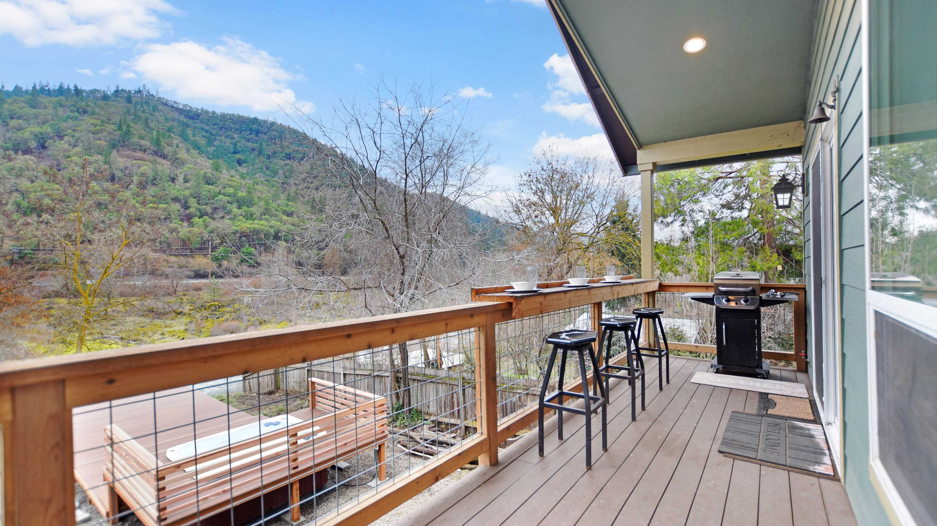 A wooden deck with a grill and bar stools, overlooking a forested hillside.