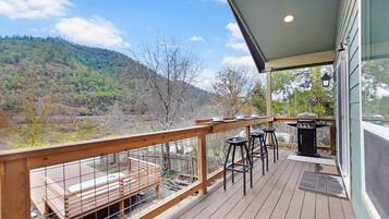 A wooden deck with a grill and bar stools, overlooking a forested hillside.