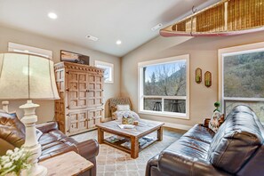 A living room with a brown leather sofa, a wooden coffee table, a wooden cabinet, and a large window with a view of trees. - The Sleepy Canoe -  A Cozy River front house near everything in Southern Oregon (Gold Hill)
