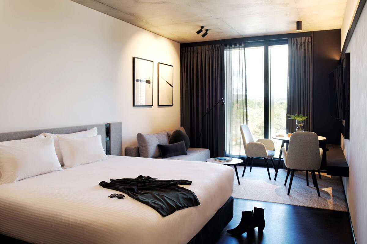 A modern hotel room with a large bed, a desk, and a chair. There are curtains, a plant, and a TV on the wall.